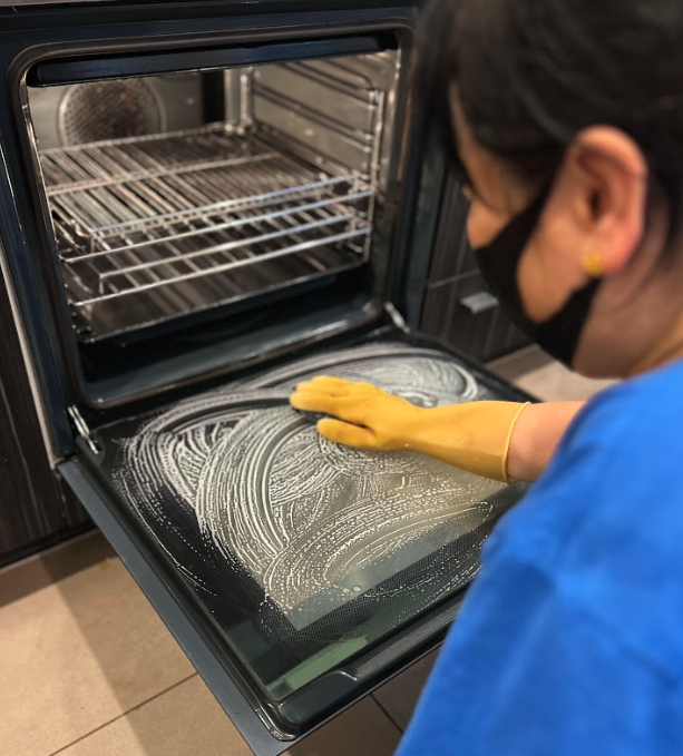 A person wearing yellow gloves scrubbing the oven door with cleaning solution.