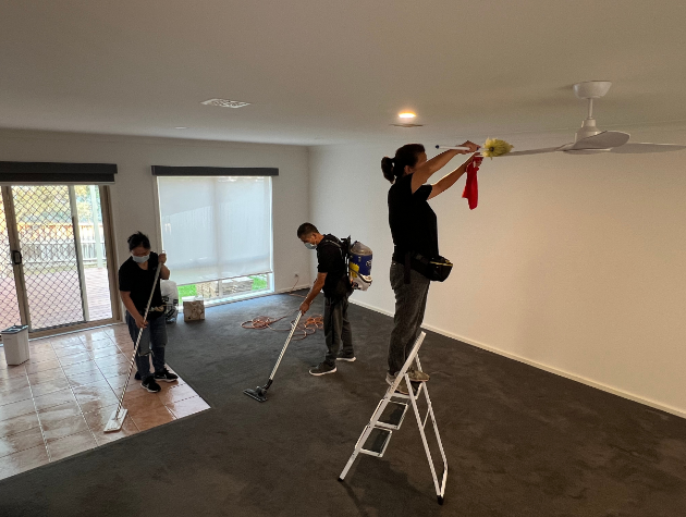 End of lease cleaning team vacuuming carpets, mopping floors, and cleaning ceiling fans in a residential living room.